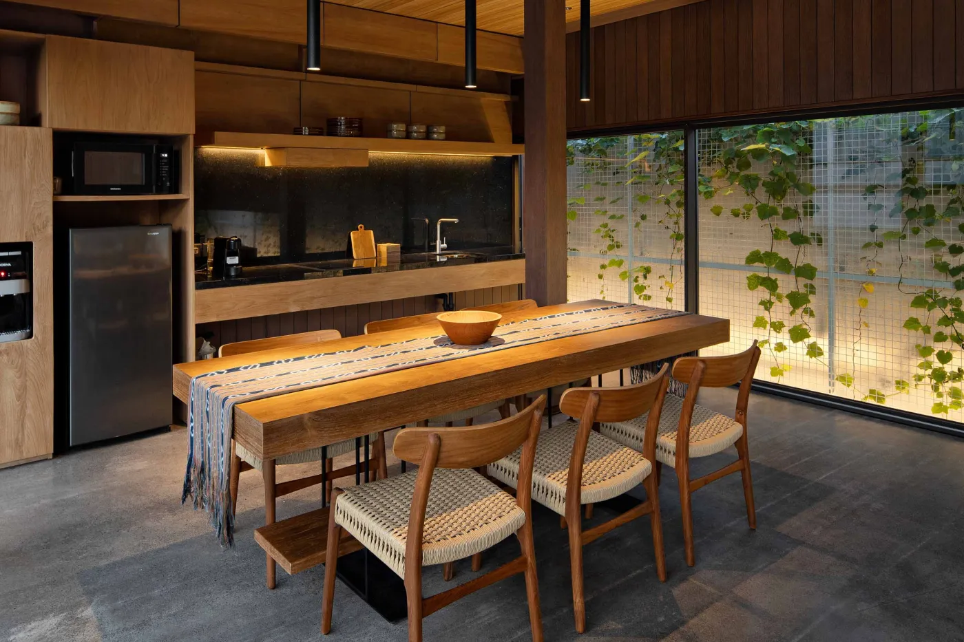 Modern wooden kitchen and dining area with pendant lights, long table set with bowl and runner, and large frosted windows overlooking greenery. - Bali Villas