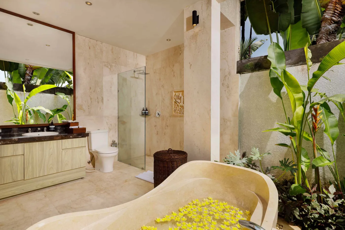 Luxurious Balinese-style bathroom featuring a petal-filled freestanding bathtub, glass shower, wooden vanity, and lush tropical plants. - Bali Villas