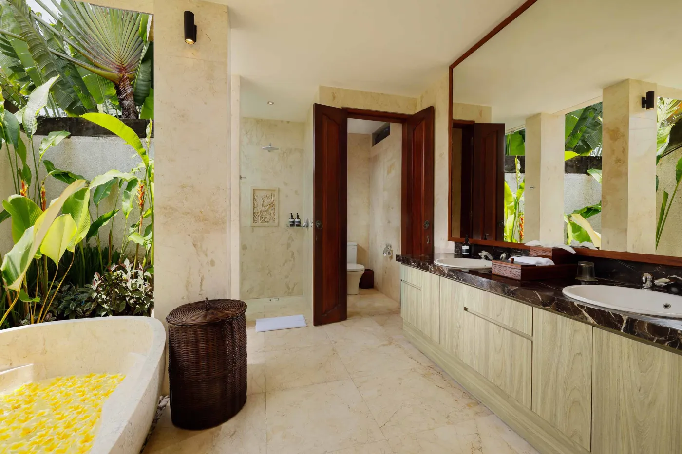 Luxurious Balinese-style bathroom featuring a petal-filled freestanding bathtub, glass shower, wooden vanity, and lush tropical plants. - Bali Villas