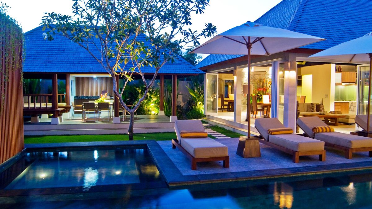 Luxurious Balinese villa at dusk with infinity pool, wooden lounge chairs, and traditional blue-roofed pavilions under frangipani trees. - Bali Villas