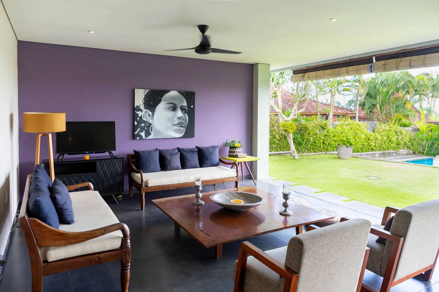 Modern Bali villa living room with purple walls, black-and-white portrait artwork, wooden sofa and coffee table, opening to lush tropical garden and pool. - Bali Villas