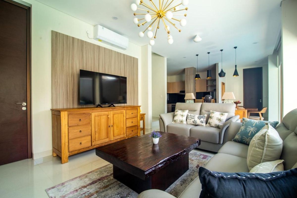 Modern open-plan living room with wooden TV cabinet, beige sofas, rustic coffee table, chandelier, and visible kitchen area. - Bali Villas