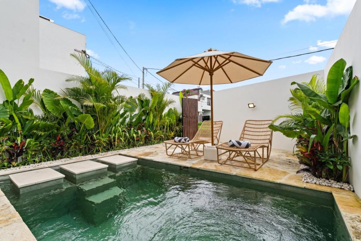 Serene private infinity pool in a tropical villa, surrounded by lush green plants, white walls, wooden lounge chairs, and a beige umbrella under a blue sky. - Bali Villas