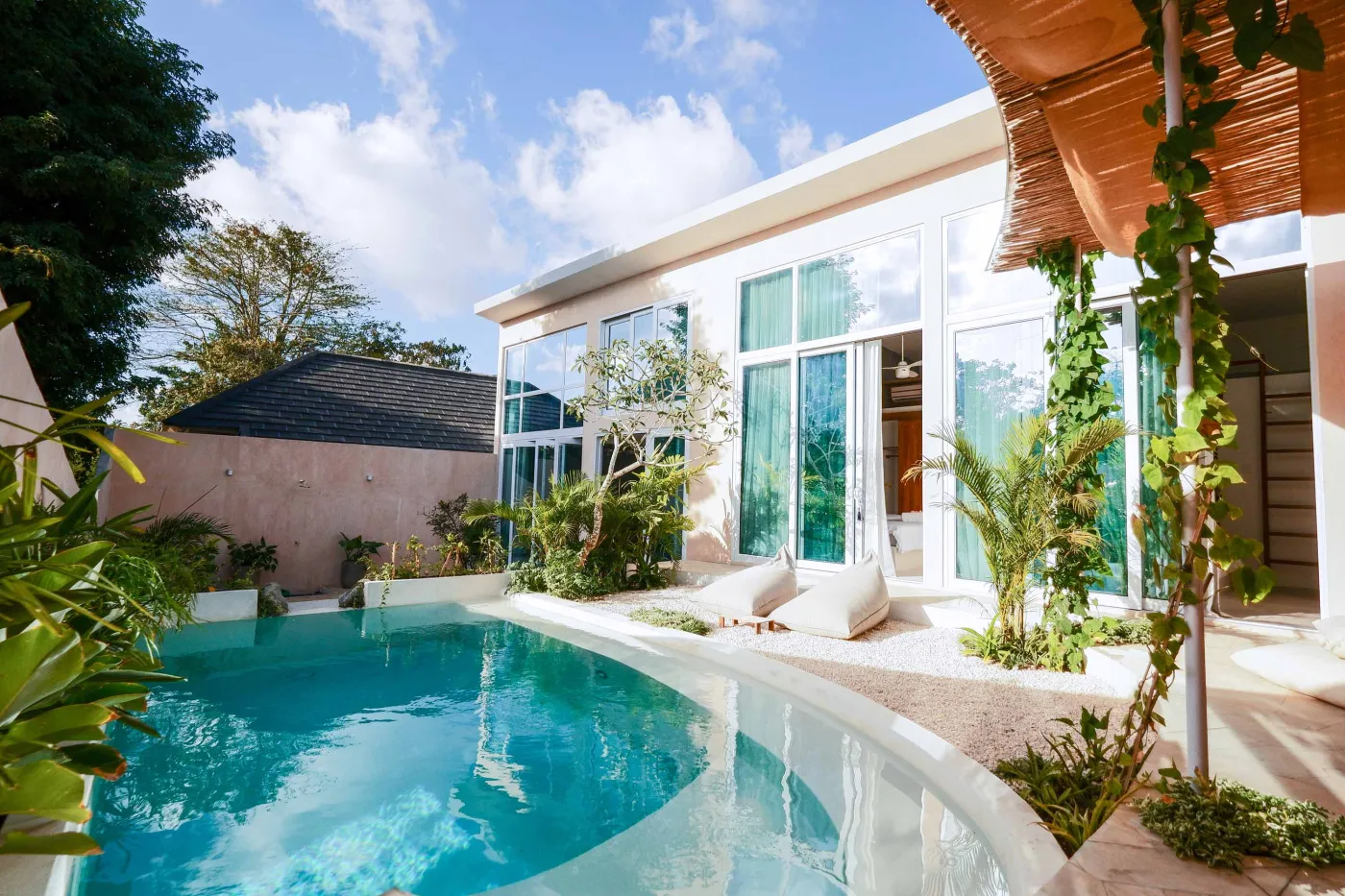 Modern tropical villa with infinity pool, lounge chairs, lush greenery, and large glass windows under a sunny sky. - Bali Villas