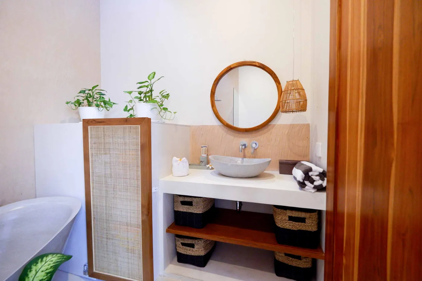 Serene bohemian bathroom with white bathtub, wooden vanity, rattan mirror, potted palm, and woven accents. - Bali Villas