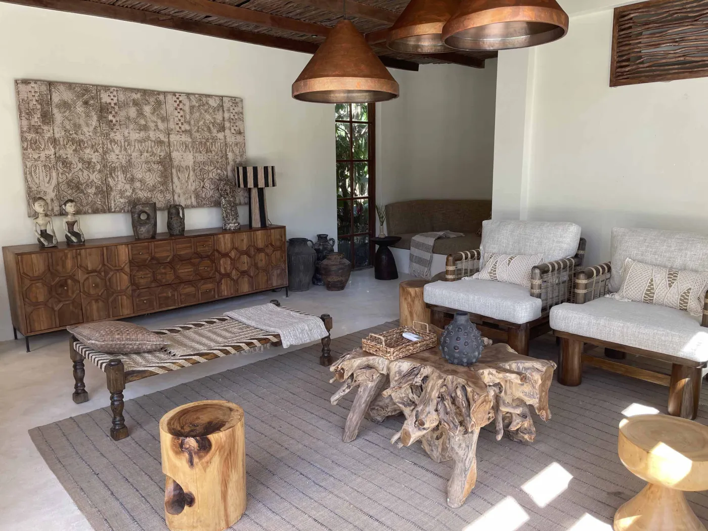 Cozy Balinese-style lounge with arched niche walls displaying pottery and sculptures, gray L-shaped sofa with patterned pillows, wooden trunk coffee table, and large window. - Bali Villas