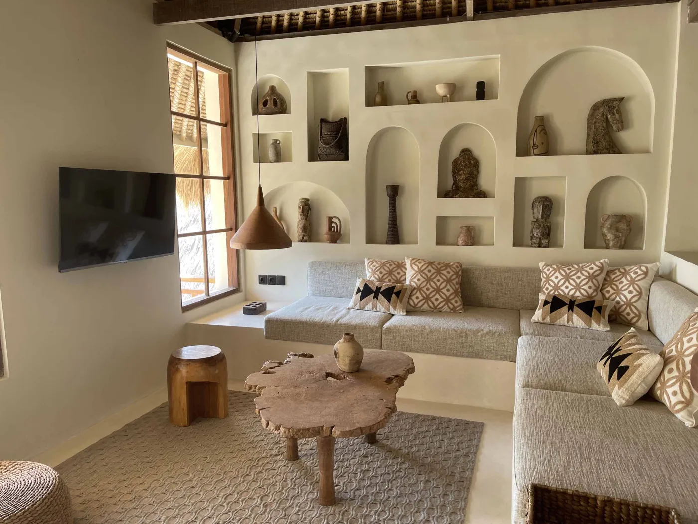 Cozy Balinese-style lounge with arched niche walls displaying pottery and sculptures, gray L-shaped sofa with patterned pillows, wooden trunk coffee table, and large window. - Bali Villas