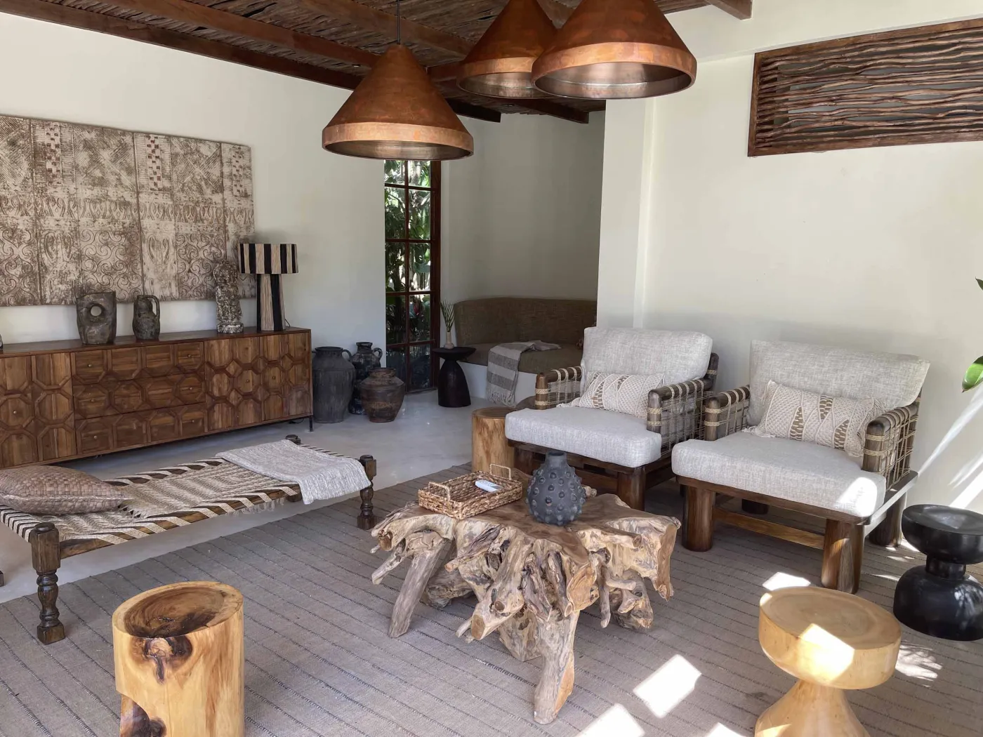 Cozy Balinese-style lounge with arched niche walls displaying pottery and sculptures, gray L-shaped sofa with patterned pillows, wooden trunk coffee table, and large window. - Bali Villas