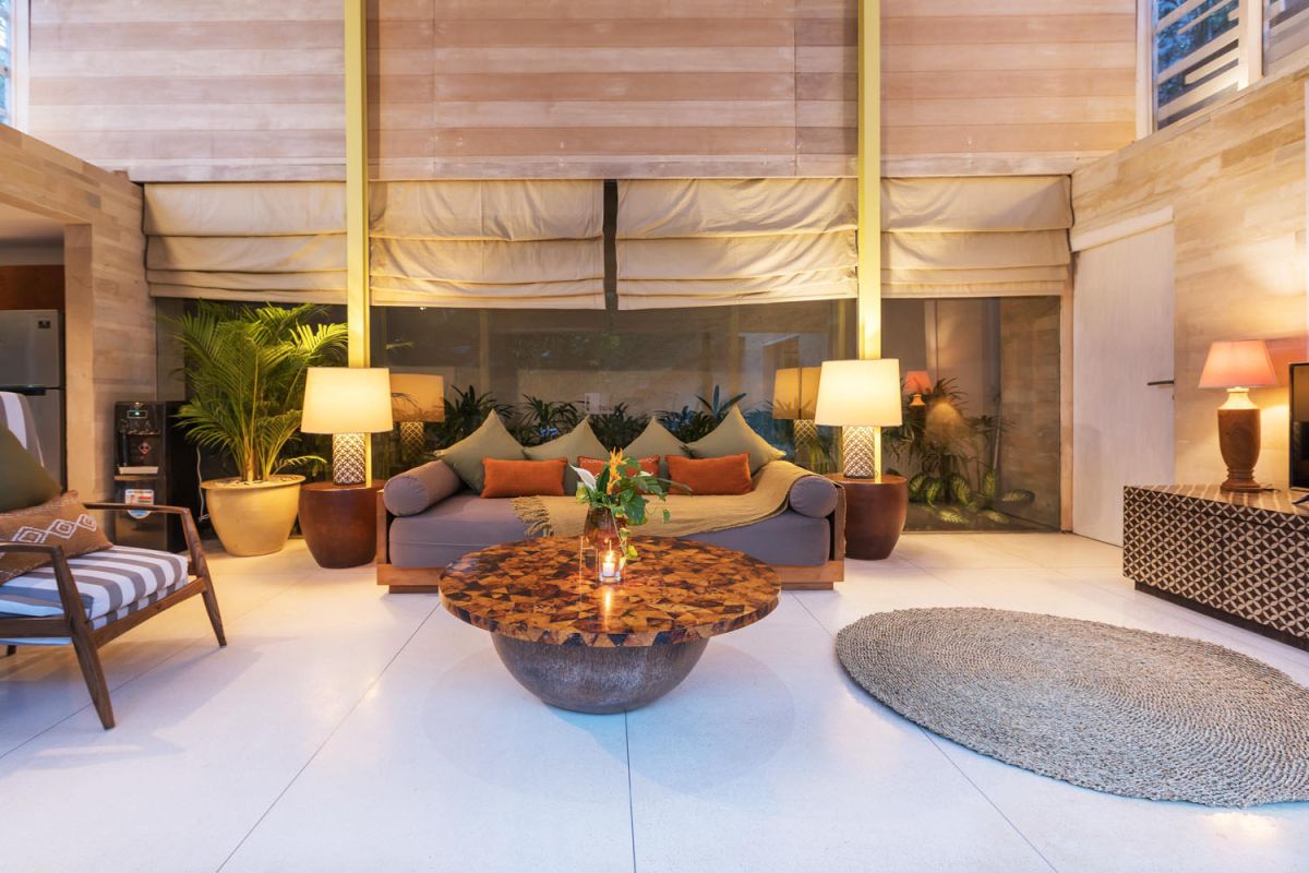 Spacious modern tropical villa interior with wooden furniture, lush plants, and open views to a pool. - Bali Villas