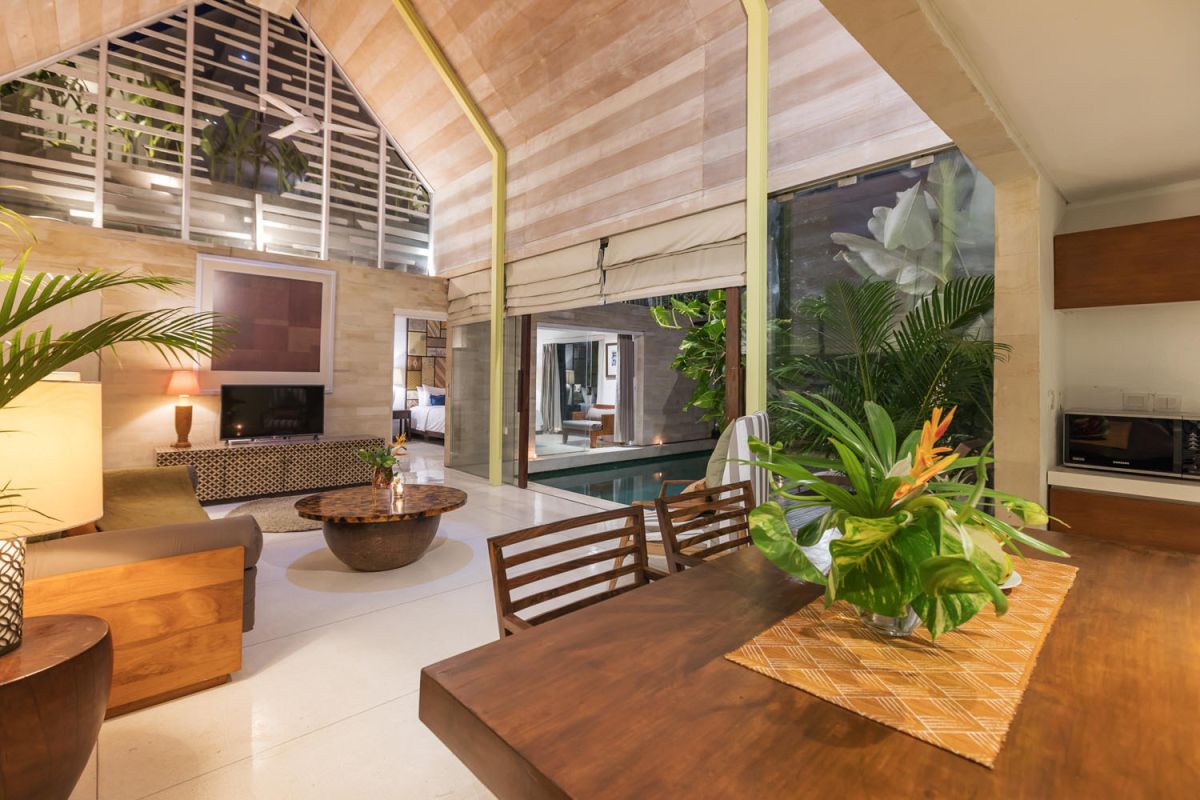 Spacious modern tropical villa interior with wooden furniture, lush plants, and open views to a pool. - Bali Villas