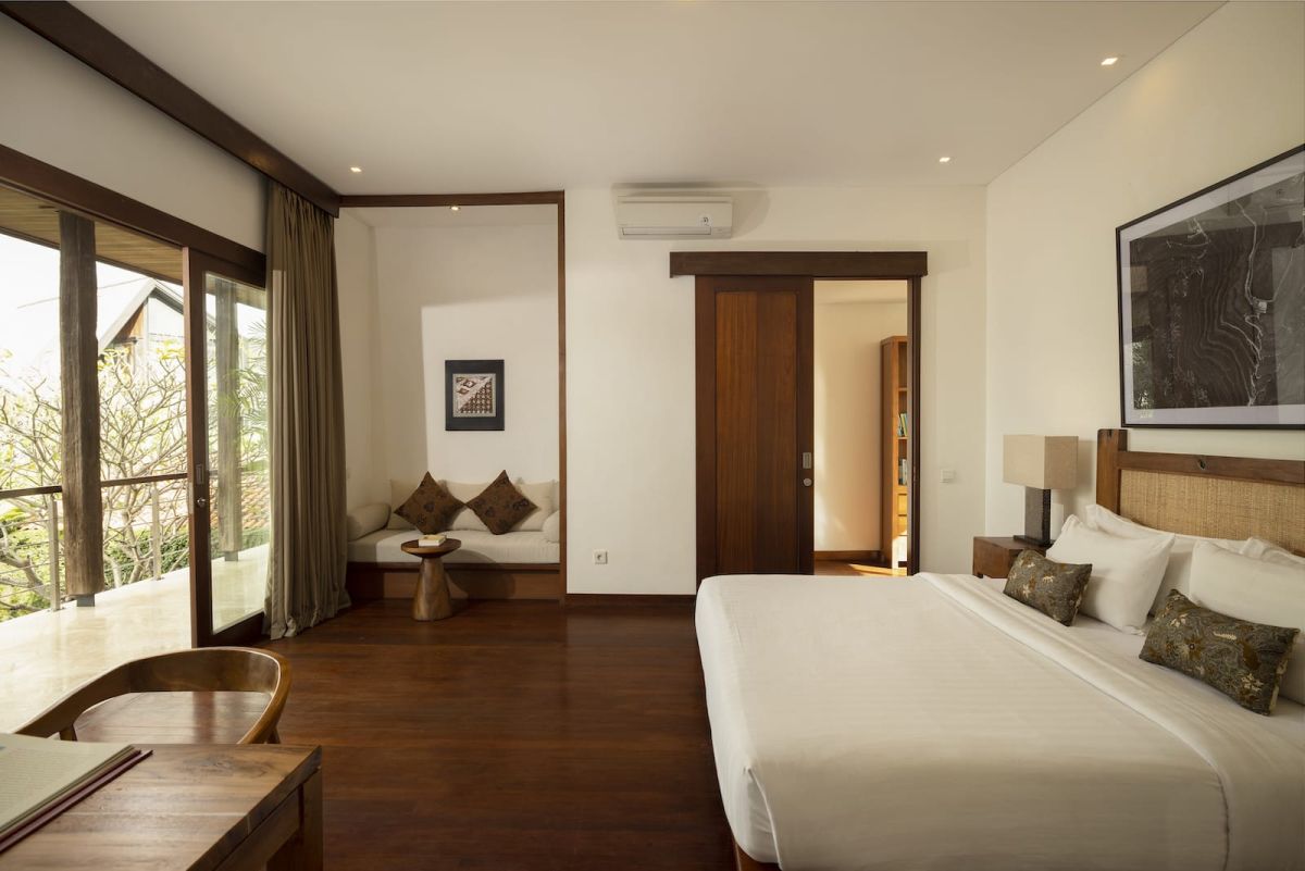 Spacious Bali villa bedroom with wooden floors, king bed, desk, lounge seating, and tropical garden views through large glass doors. - Bali Villas