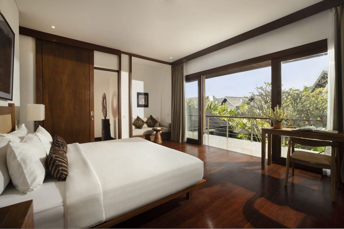 Spacious Bali villa bedroom with wooden floors, king bed, desk, lounge seating, and tropical garden views through large glass doors. - Bali Villas