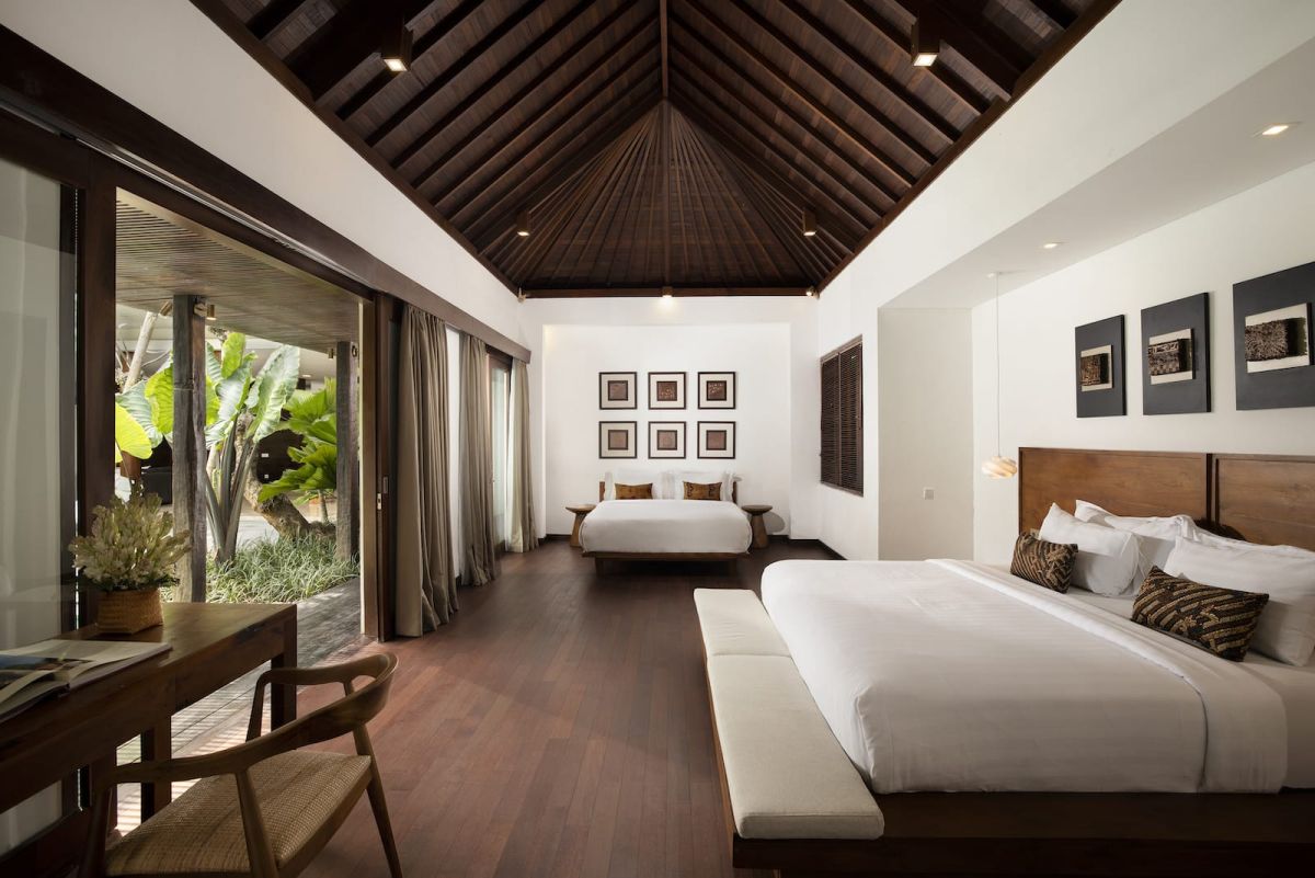 Spacious Bali villa bedroom with wooden floors, king bed, desk, lounge seating, and tropical garden views through large glass doors. - Bali Villas