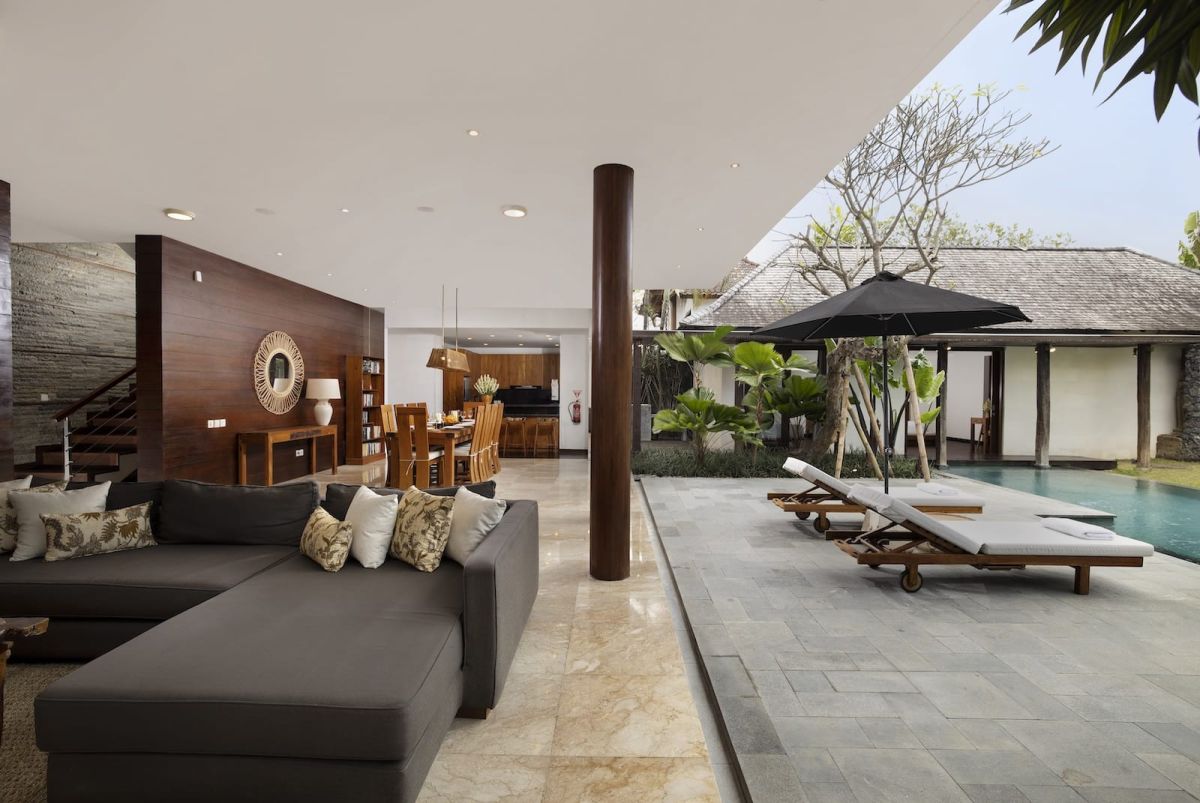 Spacious Bali villa bedroom with wooden floors, king bed, desk, lounge seating, and tropical garden views through large glass doors. - Bali Villas