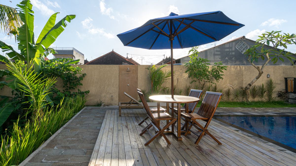 Serene Bali villa poolside patio with wooden dining set under blue umbrella, surrounded by tropical plants and traditional architecture. - Bali Villas