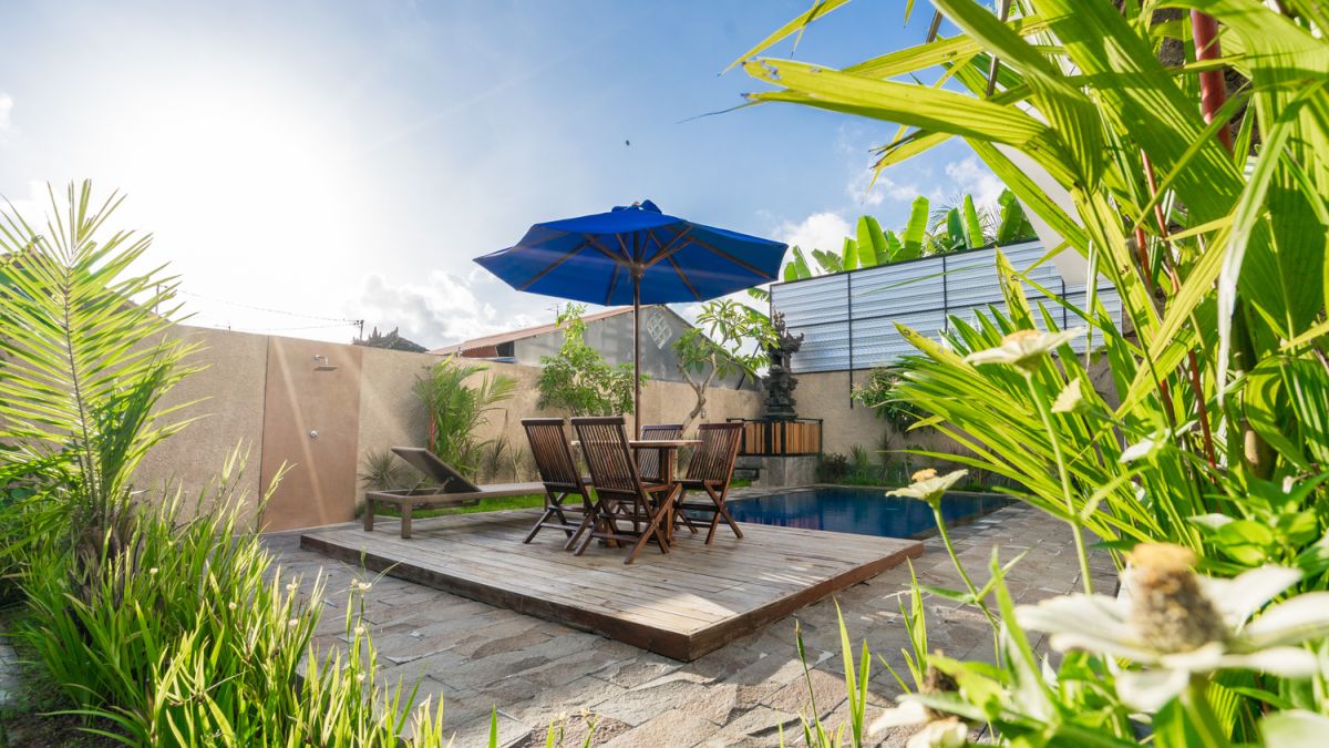 Serene Bali villa poolside patio with wooden dining set under blue umbrella, surrounded by tropical plants and traditional architecture. - Bali Villas