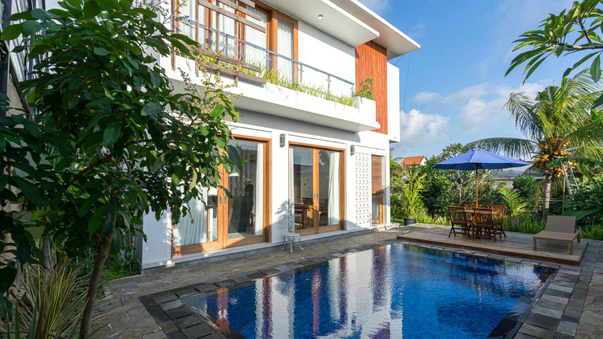 Modern tropical villa with white walls, wooden accents, large glass doors, and a serene blue infinity pool surrounded by palm trees, lounge chairs, and a blue umbrella. - Bali Villas