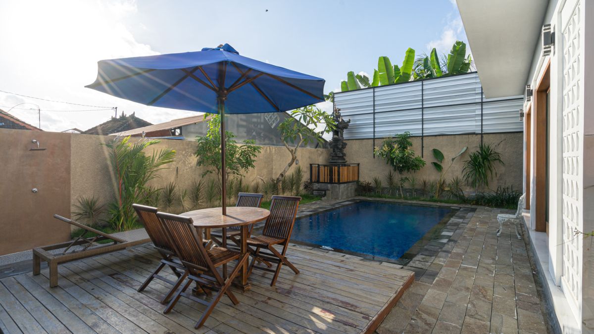 Serene Bali villa poolside patio with wooden dining set under blue umbrella, surrounded by tropical plants and traditional architecture. - Bali Villas