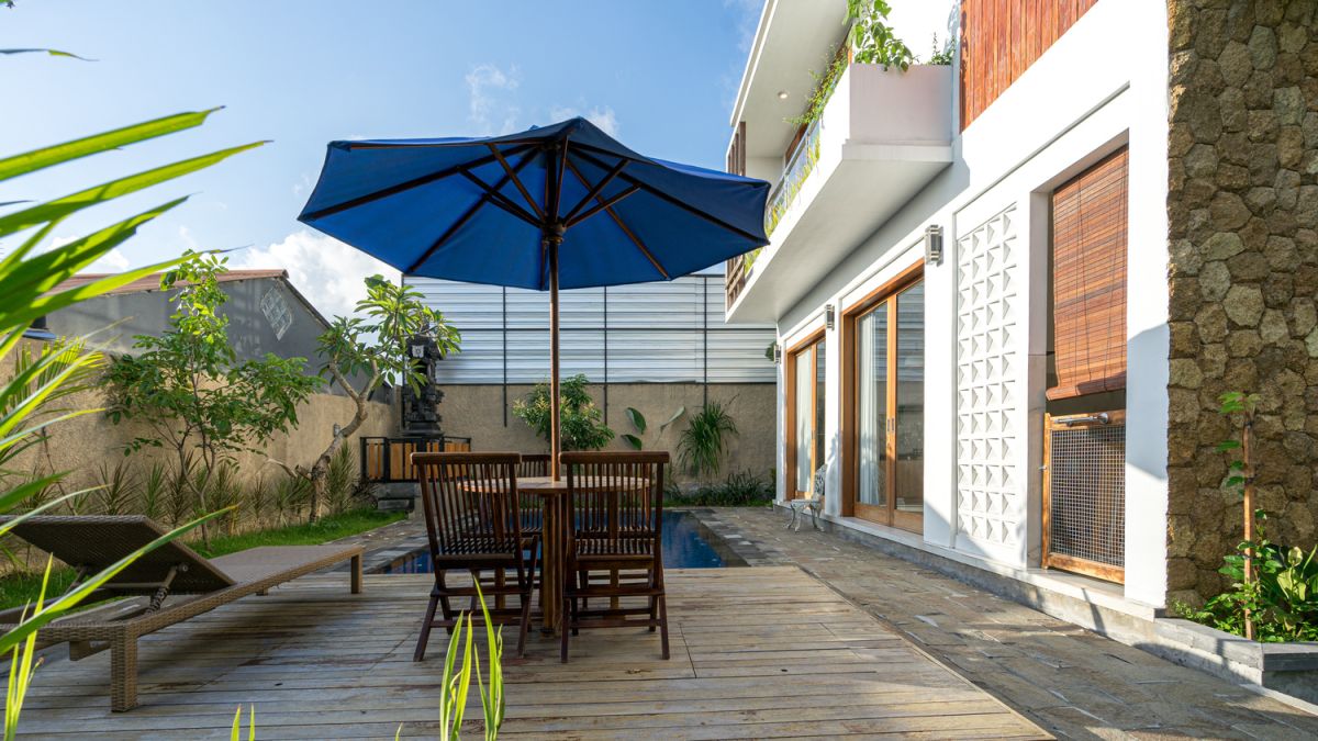 Serene Bali villa poolside patio with wooden dining set under blue umbrella, surrounded by tropical plants and traditional architecture. - Bali Villas