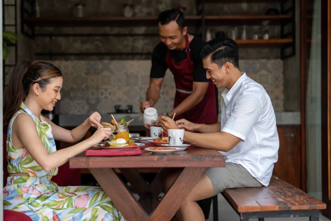Rustic tropical kitchen with wooden cabinets, shelves of jars, tiled sink, and lush green plant wall behind metal frame, plus wooden dining bench and table. - Bali Villas