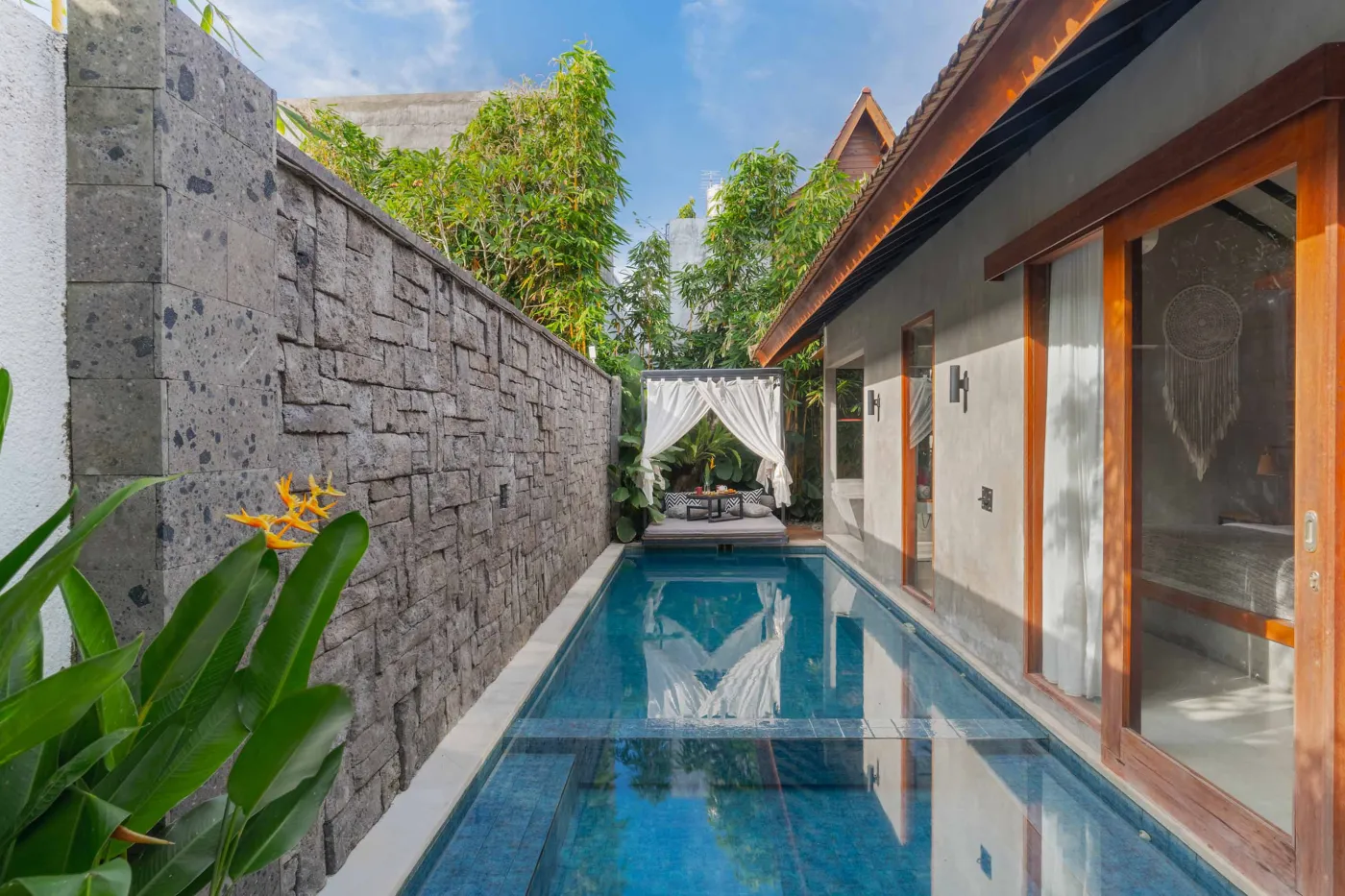Rustic tropical kitchen with wooden cabinets, shelves of jars, tiled sink, and lush green plant wall behind metal frame, plus wooden dining bench and table. - Bali Villas