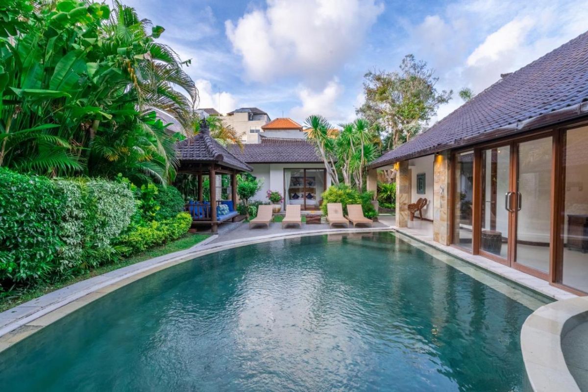 Luxurious tropical villa pool in Bali with lounge chairs, lush greenery, and traditional Balinese architecture under a clear sky. - Bali Villas