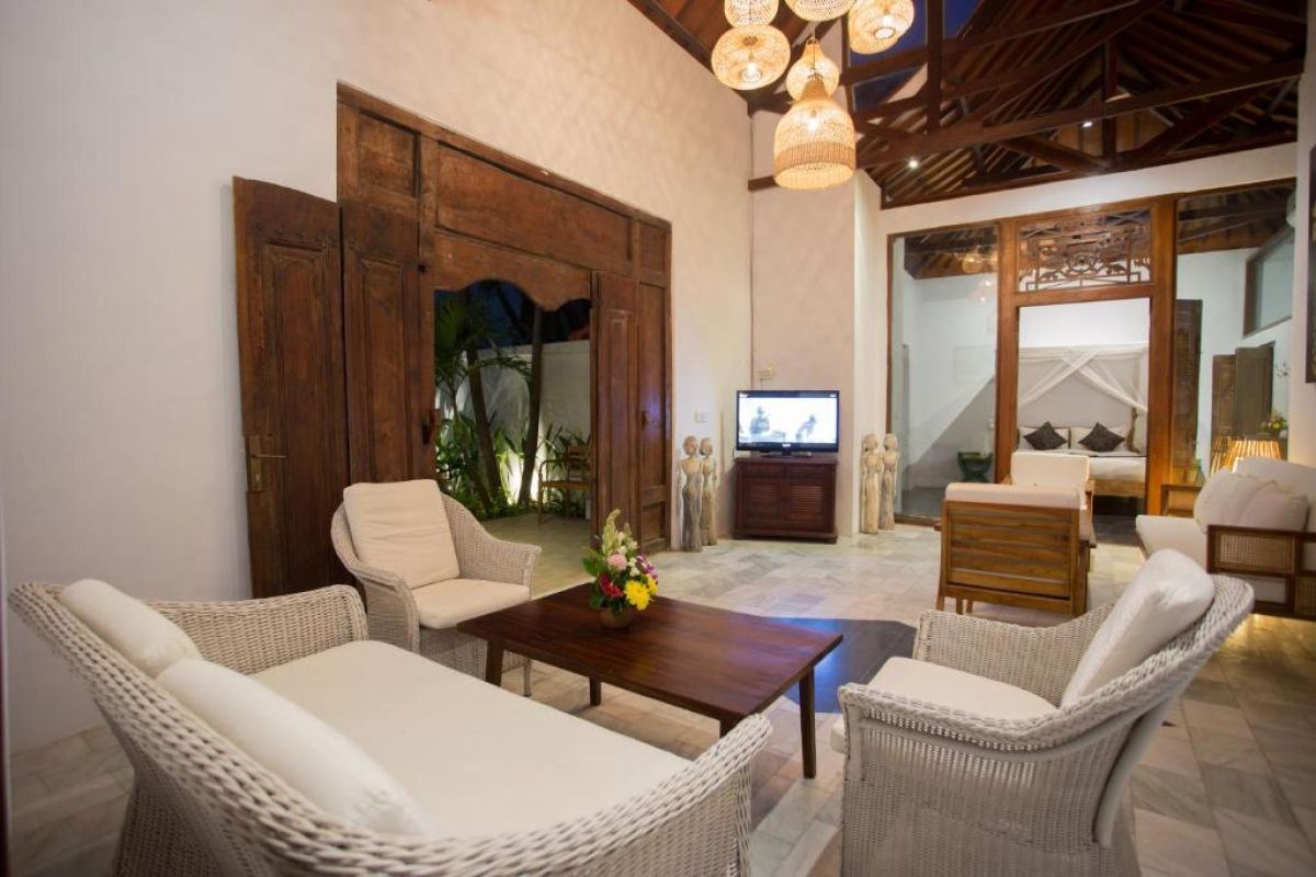 Spacious Balinese villa living area with rattan furniture, wooden accents, pendant lanterns, and open doors to a lush garden bedroom. - Bali Villas