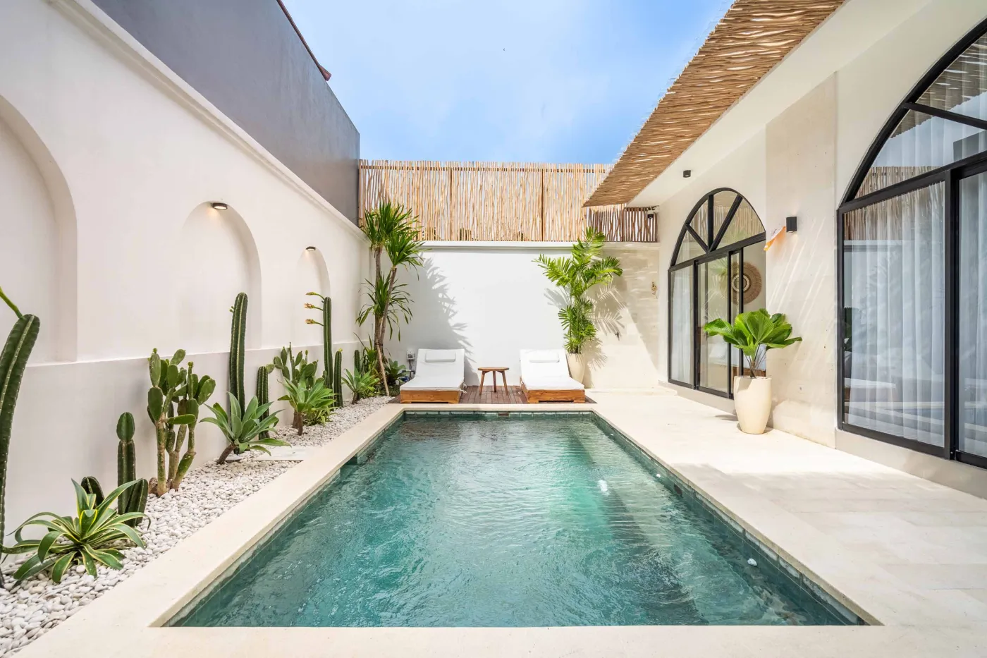 Tropical villa courtyard with infinity pool, lounge chairs, cacti, palms, and arched white architecture under a thatched roof. - Bali Villas