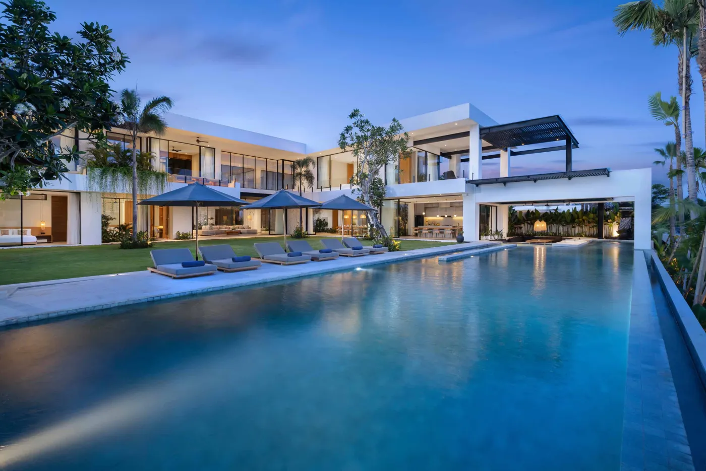 Modern tropical luxury villa at dusk, featuring an infinity pool, lounge seating, and lush palm gardens. - Bali Villas