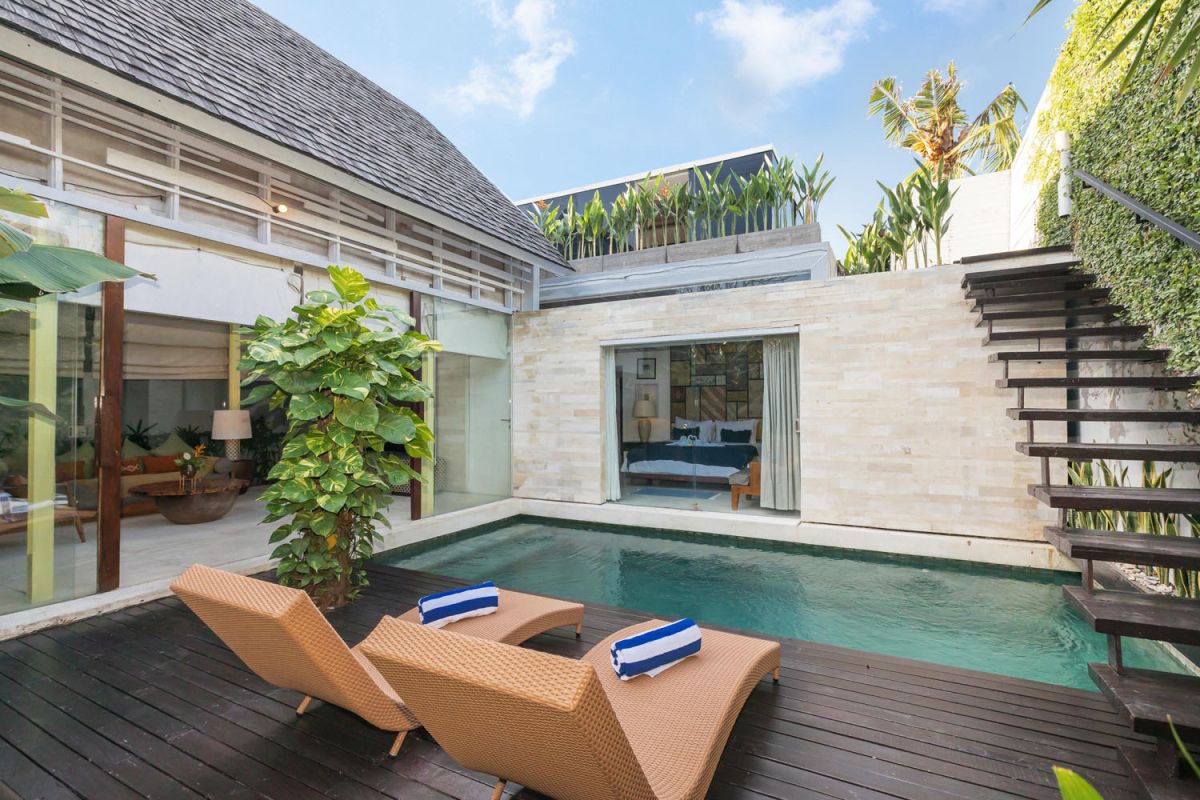 Serene private infinity pool in a modern Balinese villa, surrounded by lush greenery, wooden deck loungers, and glass doors to a bedroom. - Bali Villas