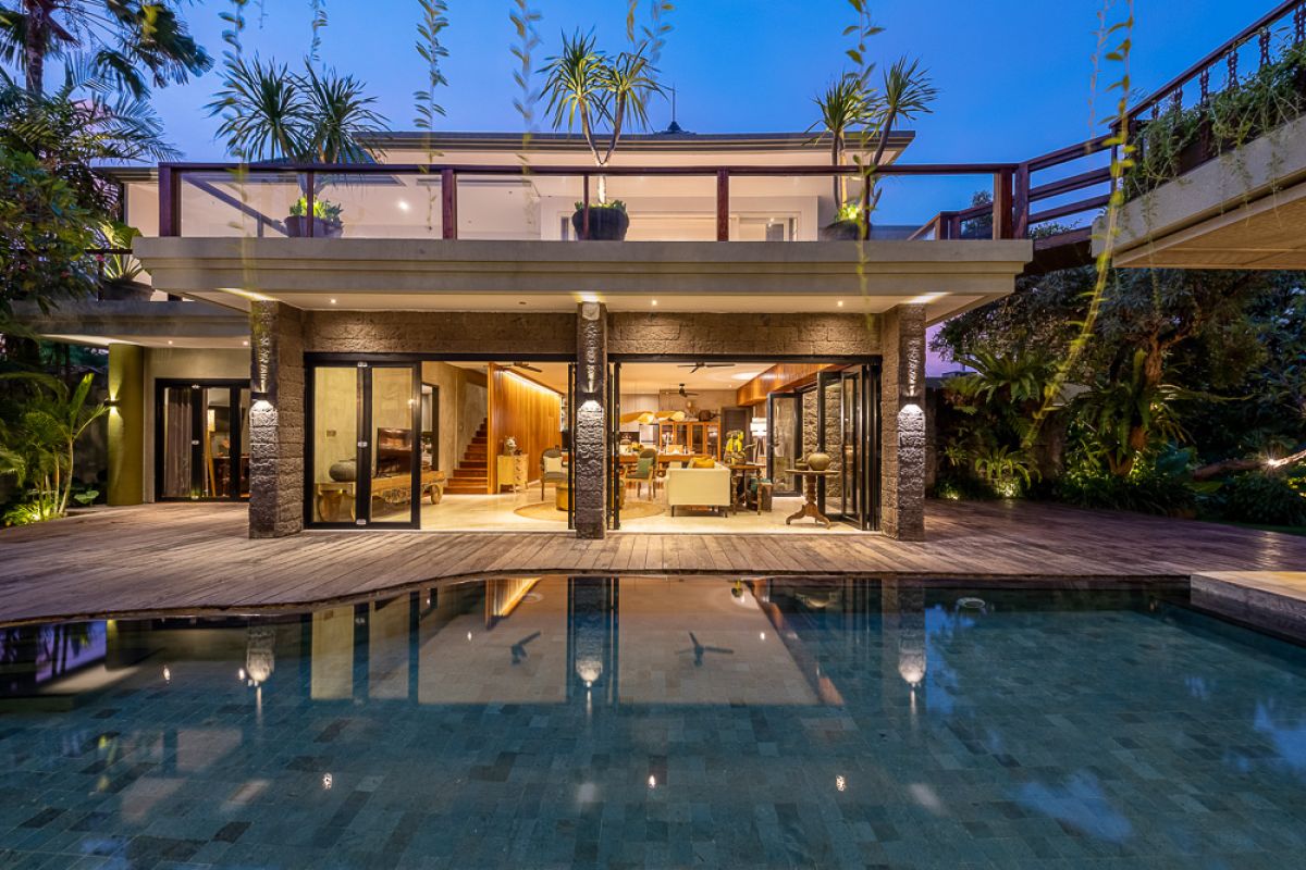 Modern tropical villa at dusk with infinity pool, wooden deck, lush greenery, and large glass doors. - Bali Villas