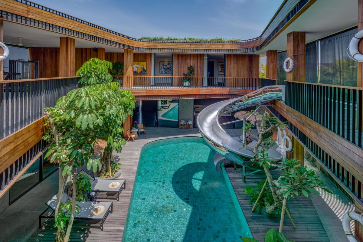 Aerial view of a lush tropical resort courtyard with a turquoise infinity pool, curving waterslide, wooden decks, and surrounding greenery. - Bali Villas
