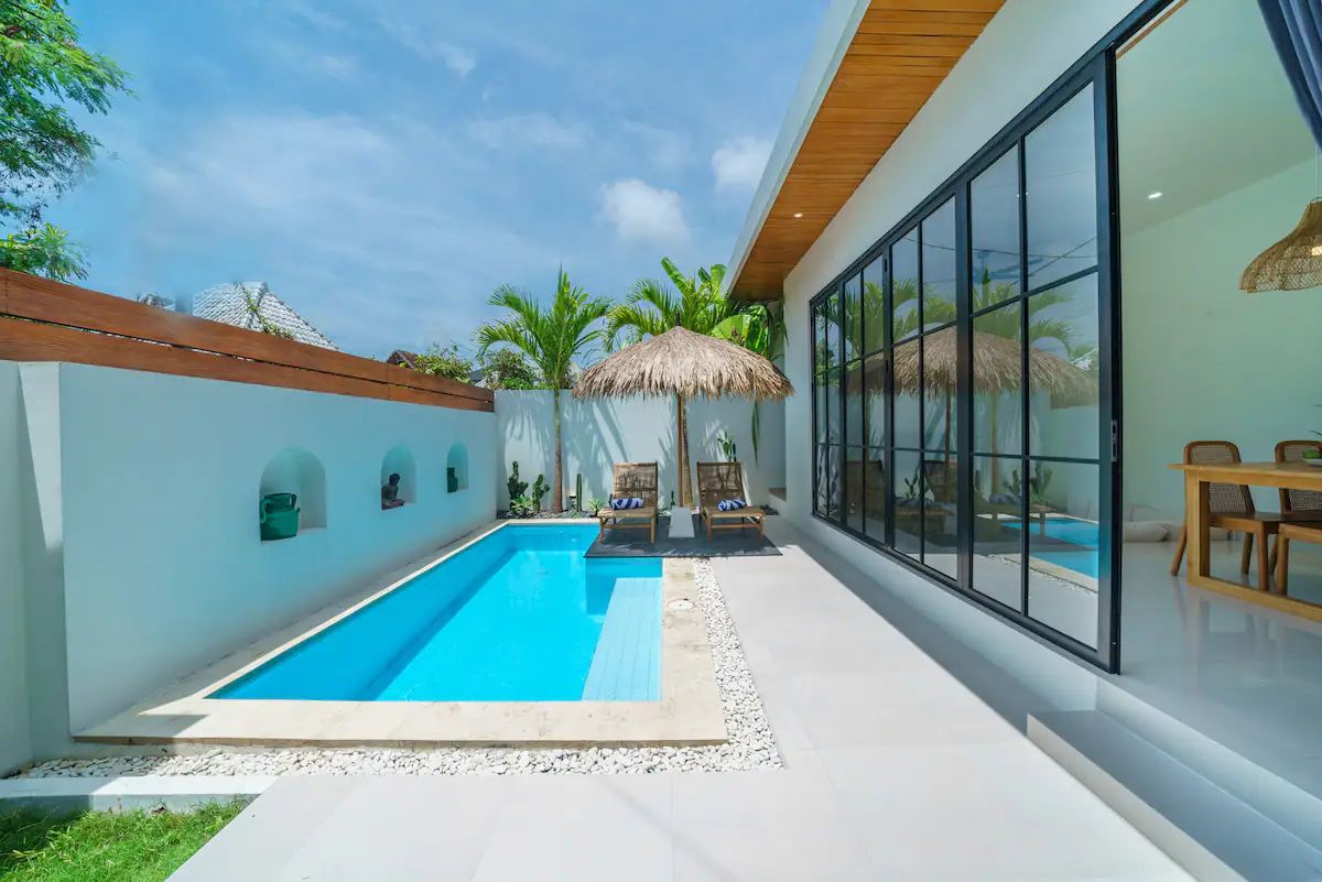 Modern tropical villa poolside with blue lap pool, wooden loungers, palm trees, and large glass windows. - Bali Villas