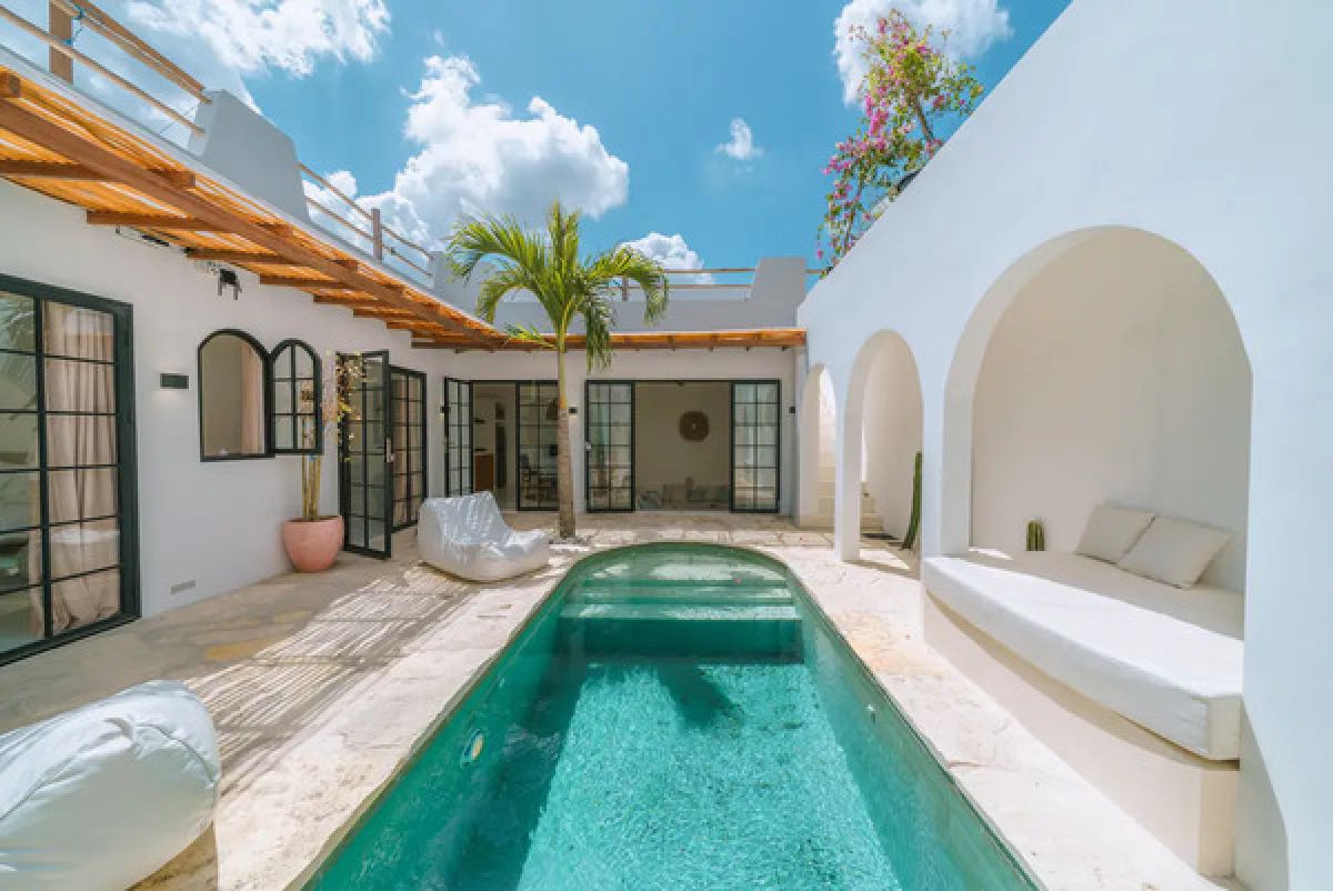 Serene tropical courtyard villa with turquoise pool, white arched walls, palm tree, and lounge seating under blue skies. - Bali Villas