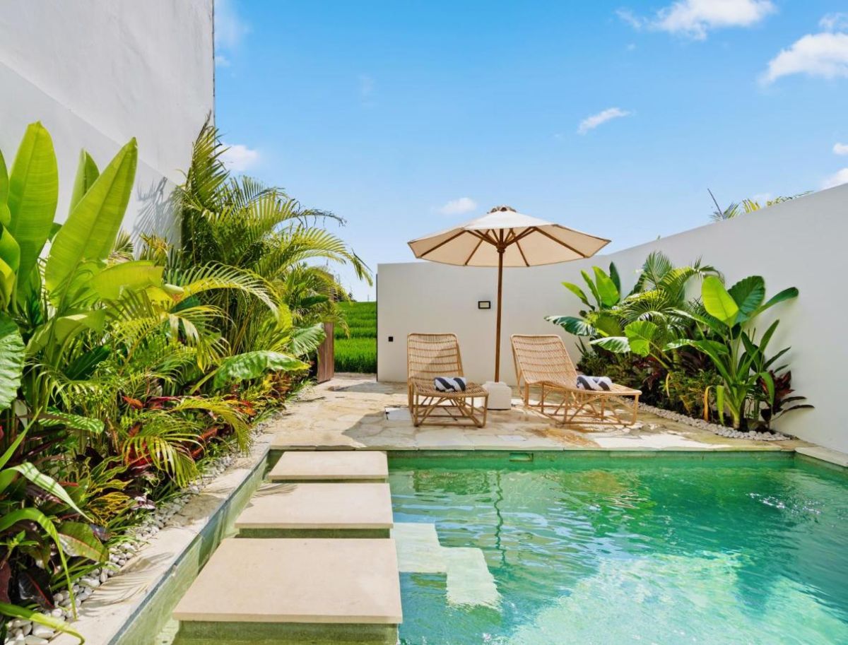 Serene tropical private pool with teal water, surrounded by lush palms and white walls, featuring wooden lounge chairs under a beige umbrella. - Bali Villas