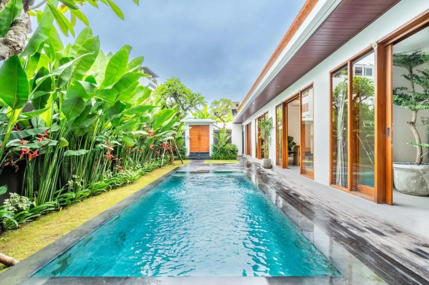 Luxurious long blue swimming pool in a modern Balinese villa, surrounded by tropical banana plants and open sliding glass doors. - Bali Villas