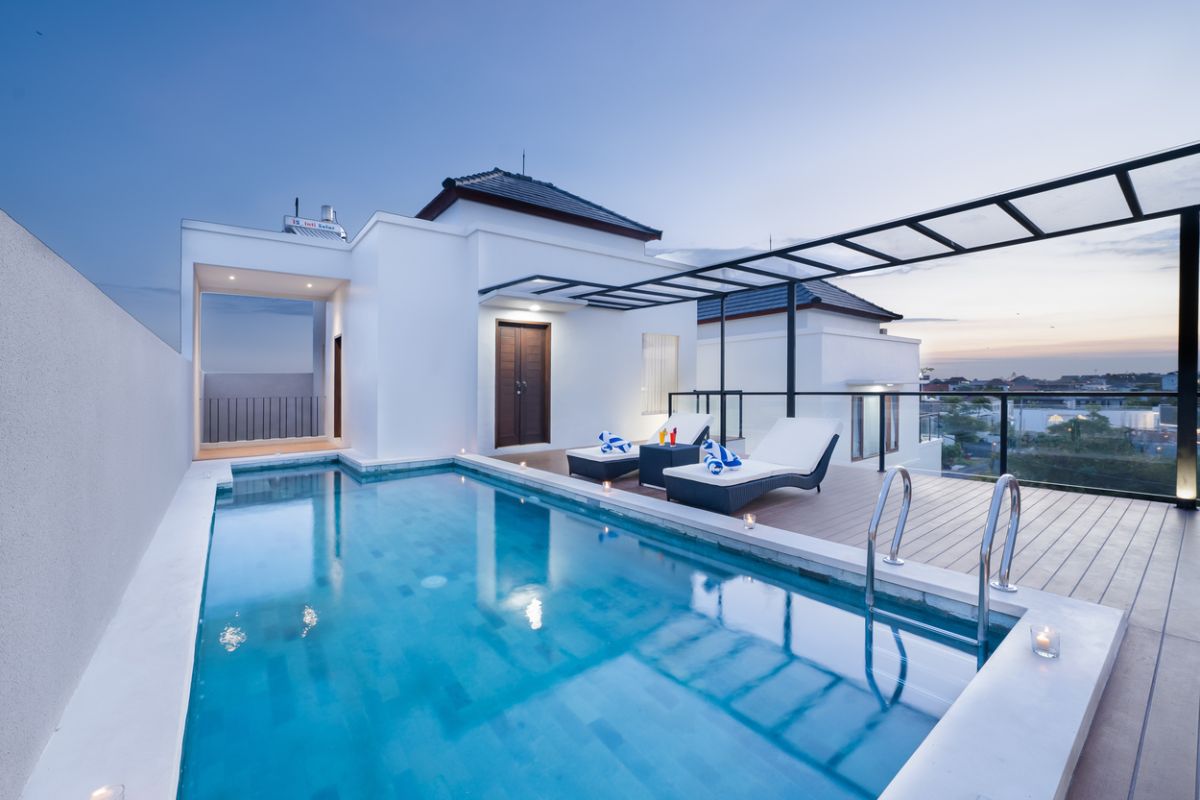 Luxurious rooftop infinity pool at sunset with white lounge chairs, blue towels, and modern deck overlooking city skyline. - Bali Villas