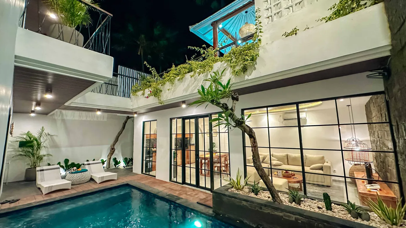 Modern tropical villa at night featuring a lit blue infinity pool, outdoor lounge seating, lush plants, and glass-walled architecture. - Bali Villas
