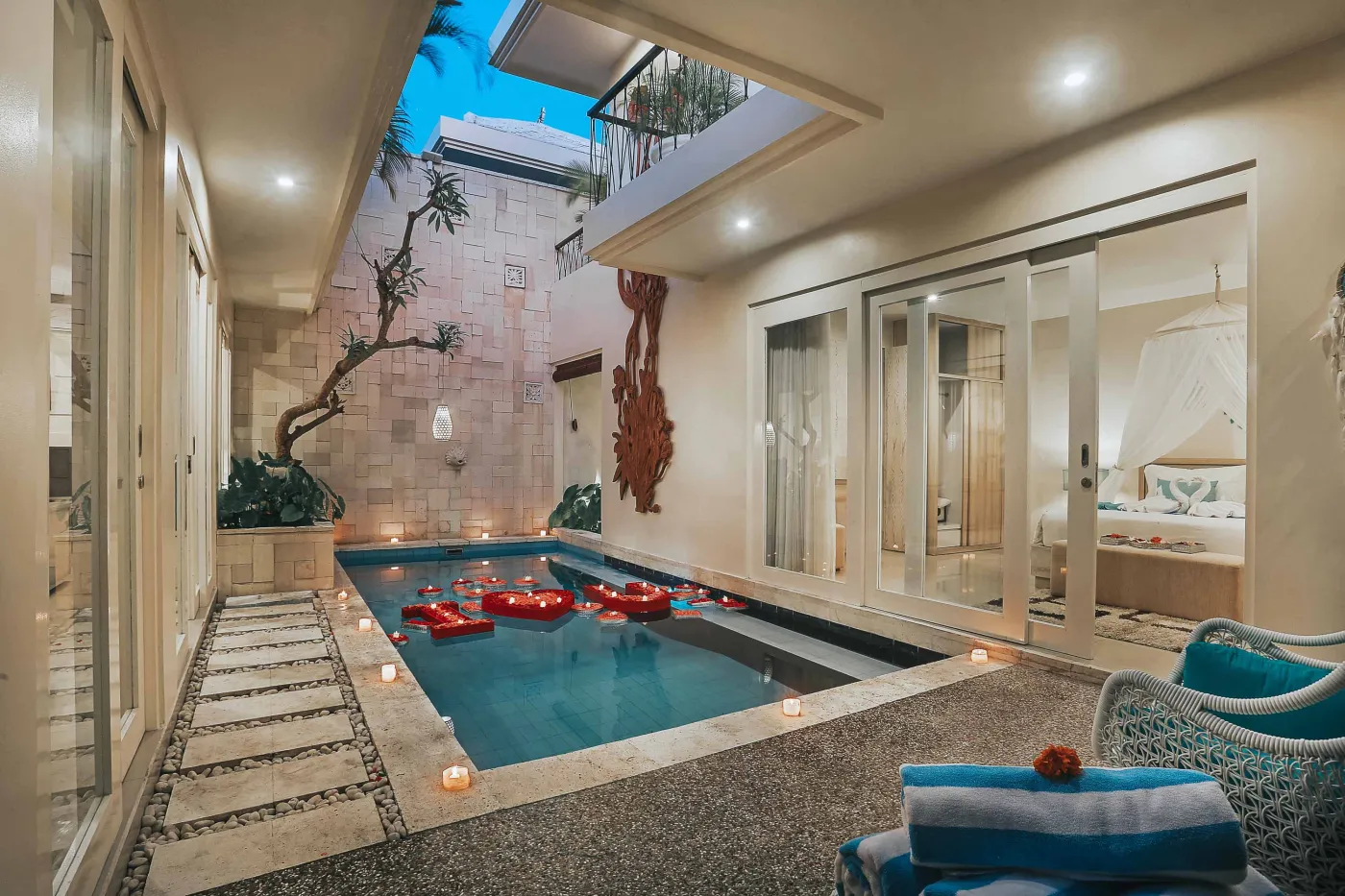 Luxurious Balinese villa courtyard with private infinity pool, red floaties, lounge chairs, and tropical stone carvings at dusk. - Bali Villas