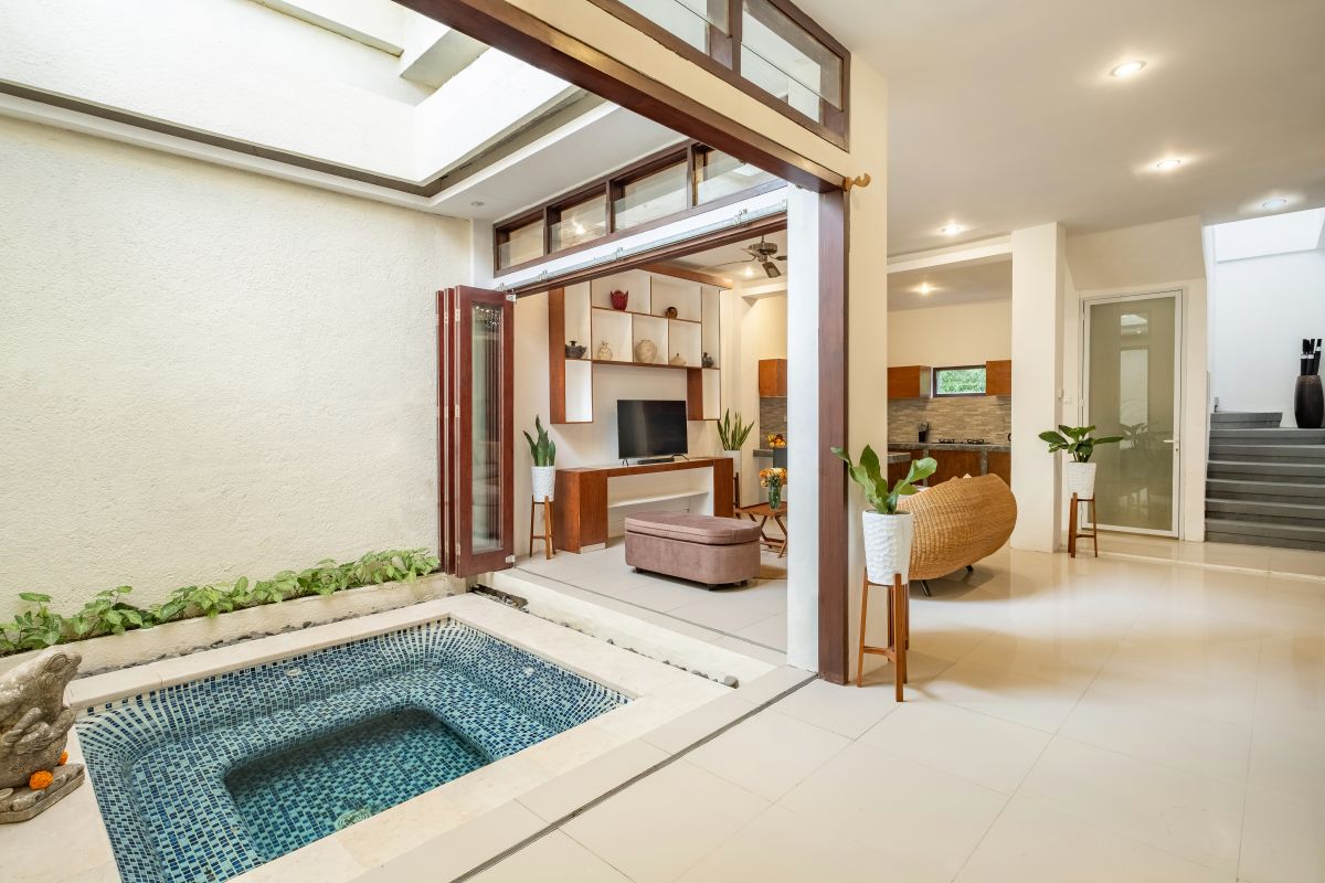 Modern tropical indoor living space with blue-tiled pool, beige sofa, potted plants, wall-mounted TV, and a cat near the water. - Bali Villas