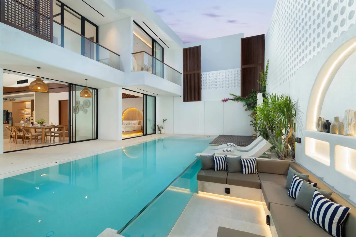 Modern white luxury villa with infinity pool, tropical plants, and cushioned outdoor lounge at sunset. - Bali Villas