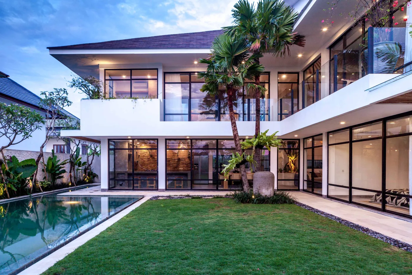 Modern tropical villa with infinity pool, palm trees, lush lawn, and expansive glass walls at dusk. - Bali Villas