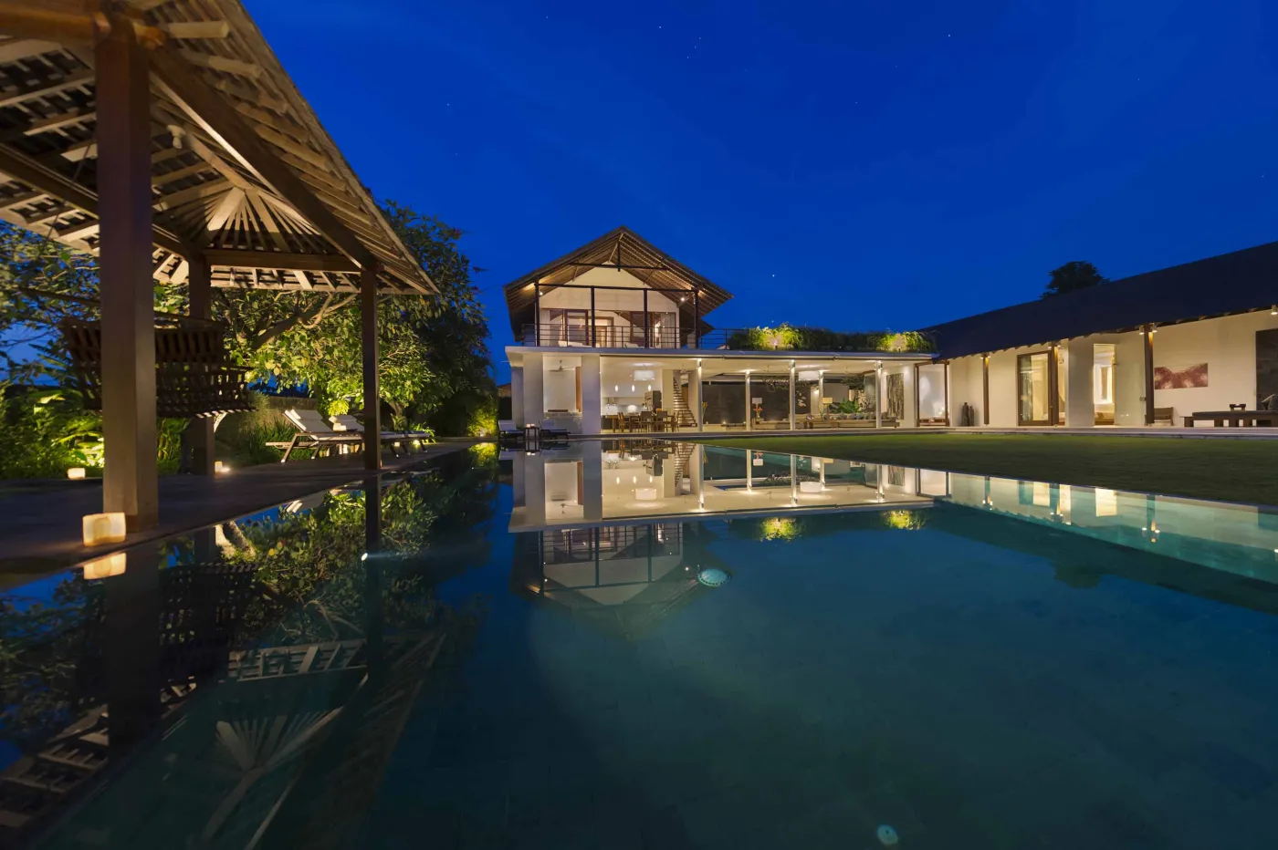 Luxurious modern tropical villa at night, reflected in an infinity pool with surrounding lanterns and greenery. - Bali Villas