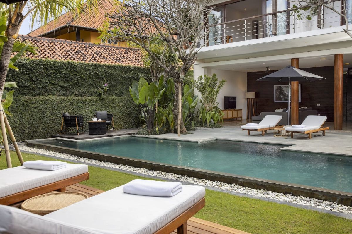 Luxurious Balinese villa poolside with turquoise infinity pool, white lounge chairs, tropical plants, and red-tiled architecture. - Bali Villas
