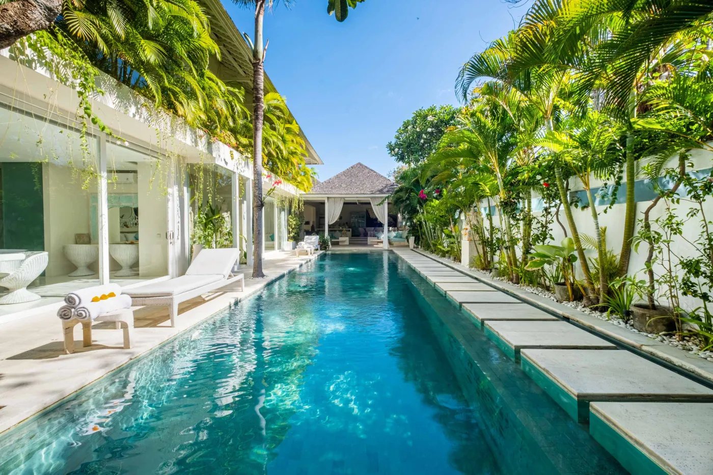Luxurious tropical infinity pool in a modern Bali-style villa, surrounded by palm trees and lush greenery under a blue sky. - Bali Villas