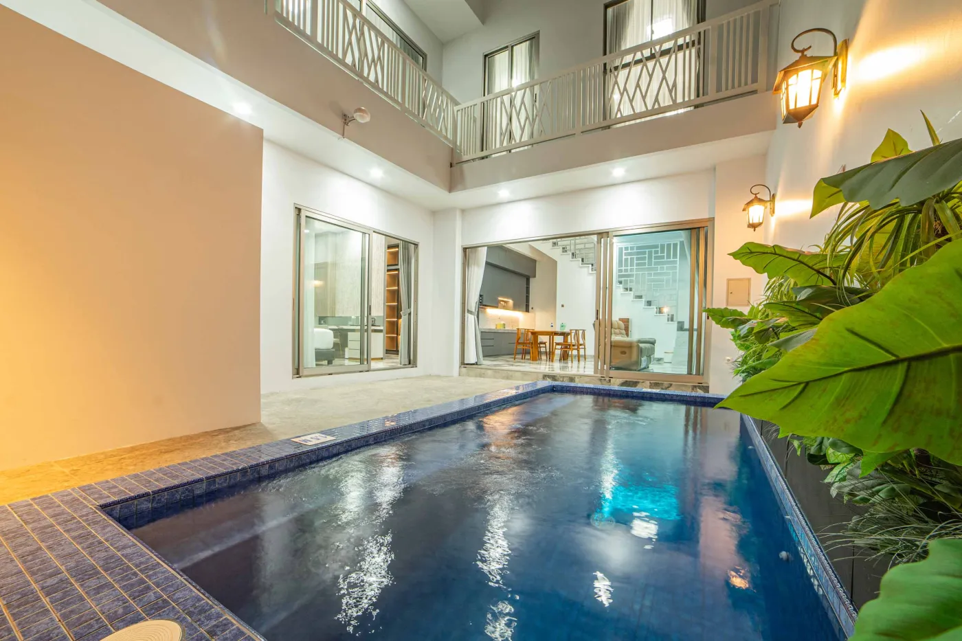 Modern indoor pool in a luxurious tropical home, featuring blue-tiled water, glass walls, dining area, balcony, and lush plants under warm lighting. - Bali Villas