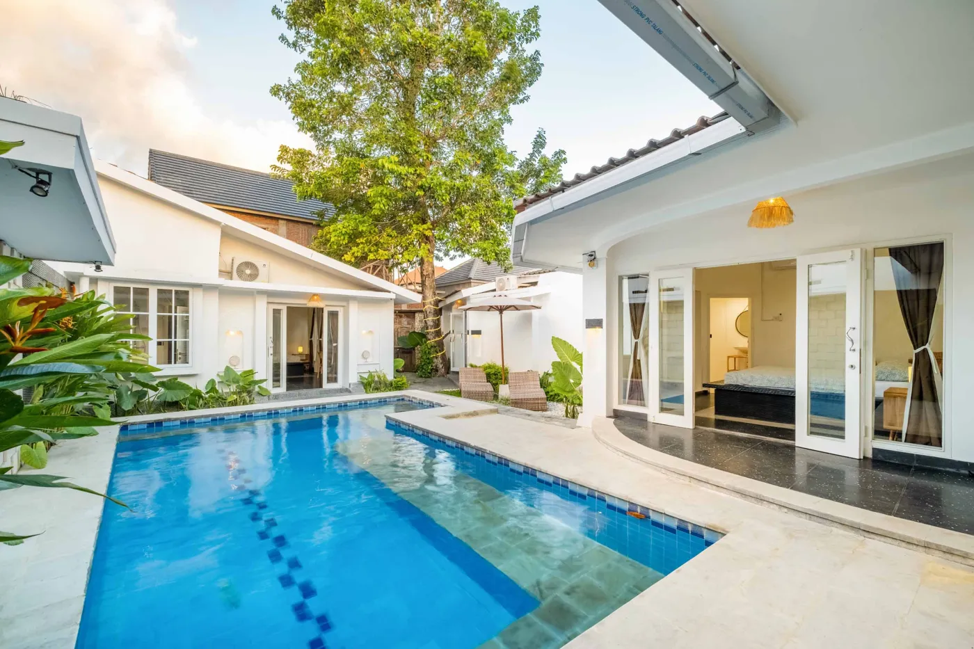 Serene Bali villa infinity pool surrounded by white architecture, lush tropical plants, and open bedroom suites at dusk. - Bali Villas