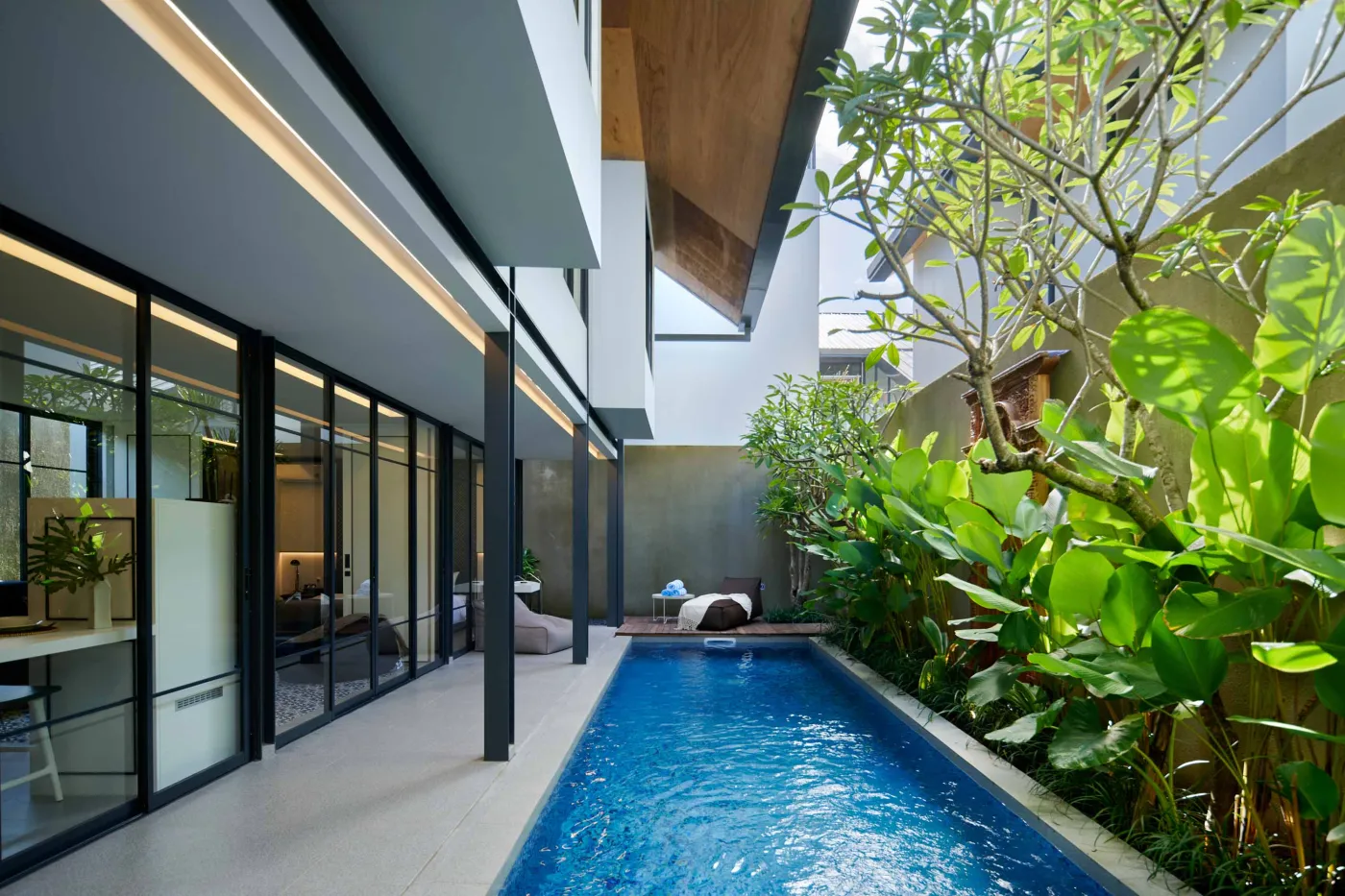 Modern tropical courtyard with a serene blue infinity pool, lush green plants, and sleek glass-and-wood architecture. - Bali Villas