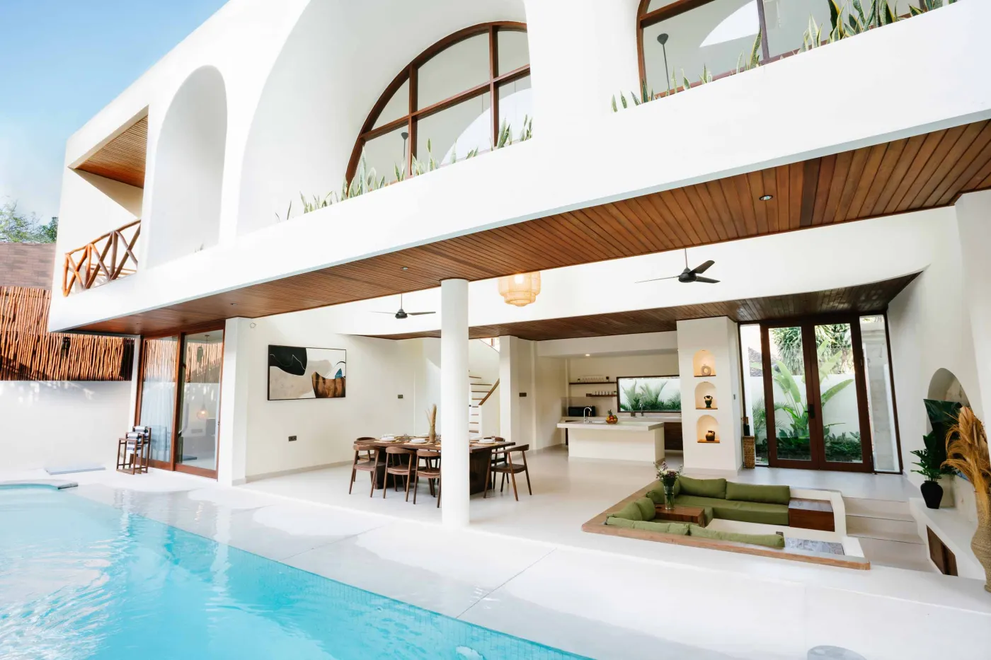 Modern tropical villa with white arched architecture, open-plan kitchen and lounge, wooden accents, green seating, and infinity pool. - Bali Villas