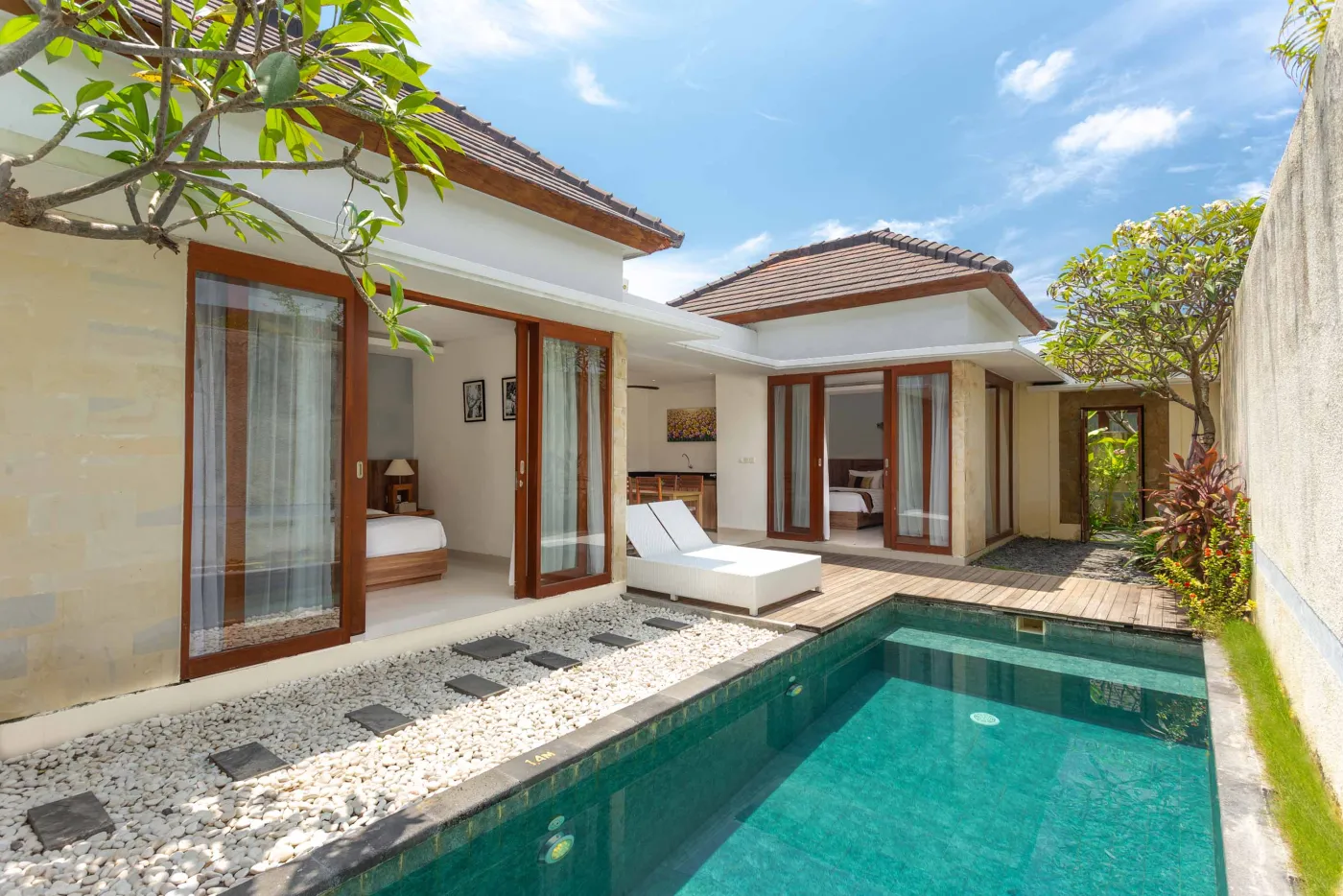 Luxurious Balinese villa with private turquoise infinity pool, wooden deck, white lounge chair, and tropical garden under blue skies. - Bali Villas